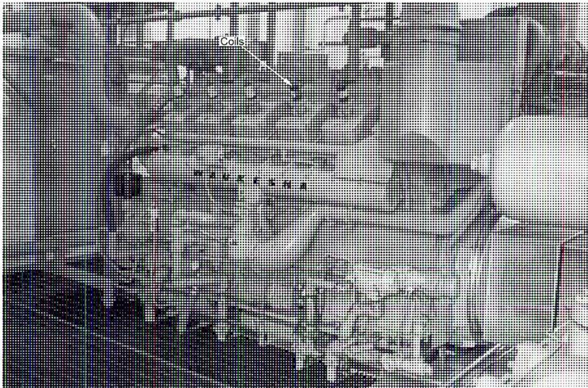 A black and white photograph of a large, industrial 12-cylinder natural gas lean burn engine. The engine is shown from a three-quarter front view, revealing its complex mechanical structure. A label 'Coils' with a leader line points to a series of spark plug coils mounted on top of the engine's intake manifold. The engine is mounted on a dark, possibly concrete, base. The background is slightly blurred, showing other industrial equipment.