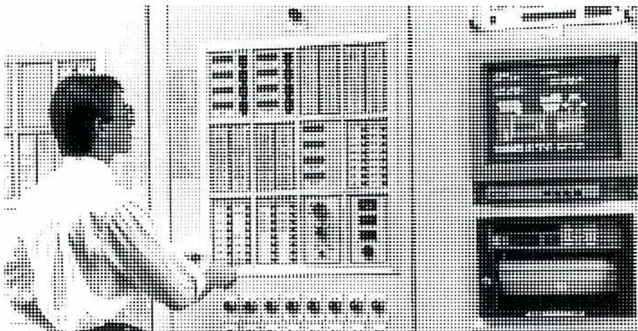 A black and white photograph showing an operator in a white shirt standing in front of a large control panel. The panel is filled with numerous dials, switches, and indicator lights. To the right of the panel are two large, vertically stacked CRT computer monitors displaying graphical process information. The operator is looking at the panel and appears to be interacting with it.