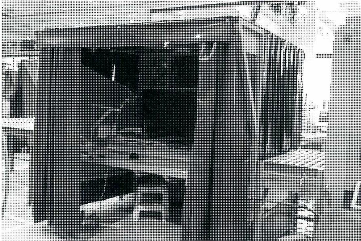 A black and white photograph of an ultraviolet inspection booth. The booth is a dark, enclosed structure with a large, dark, rectangular opening in the center, likely for the inspection area. The surrounding environment appears to be an industrial or workshop setting with various equipment and structures visible in the background.