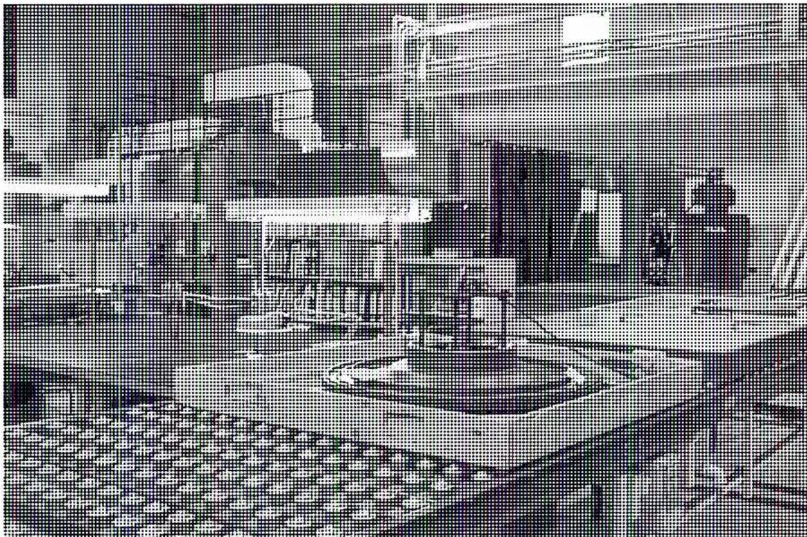 A black and white photograph showing a large industrial machine, likely a printing press, with a complex mechanical structure. The machine is situated in a factory or workshop environment. The foreground shows a large, dark, textured surface, possibly a conveyor belt or a large wheel. The background features various mechanical components, including pipes, valves, and structural elements. The lighting is bright, casting shadows on the floor.