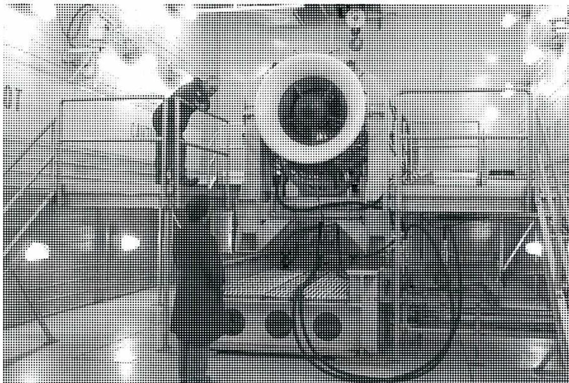 A halftone photograph of a large industrial engine, likely a gas turbine, mounted on a test stand within a specialized test cell. The engine's intake is prominent in the center, and various support structures, piping, and wiring are visible around it.