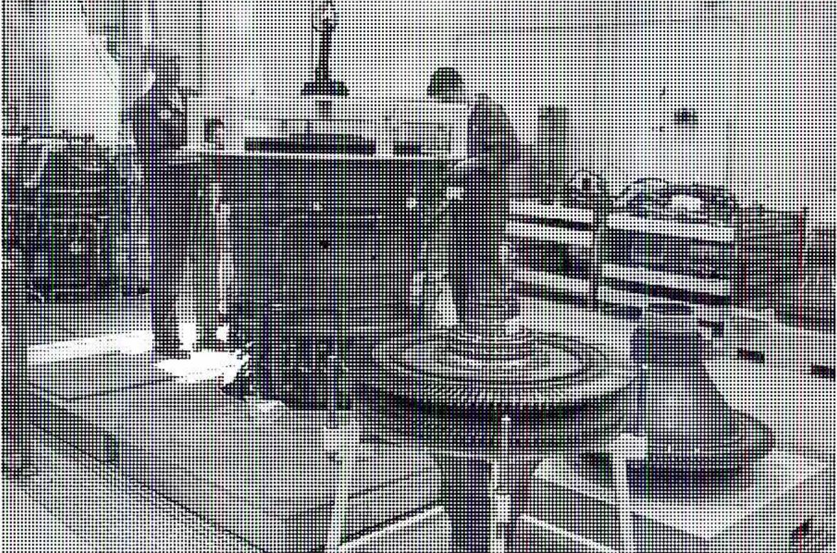 A black and white photograph showing the final reassembly of a large industrial engine. The engine is a complex, dark-colored machine with various components visible, including a large cylindrical casing and various pipes and fittings. The engine is mounted on a light-colored floor, and the background shows a workshop or factory environment with some equipment and structures visible.