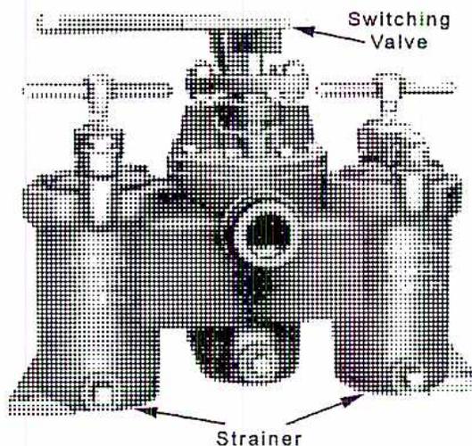 A black and white photograph of a Kraissl Duplex Strainer. The device is a complex metal assembly with two parallel strainer bodies. A central 'Switching Valve' is visible at the top, which can be rotated to direct the flow of oil into either of the two strainers. The strainer bodies have various pipes, flanges, and a central circular opening. Arrows point from the labels 'Switching Valve' and 'Strainer' to their respective parts in the image.