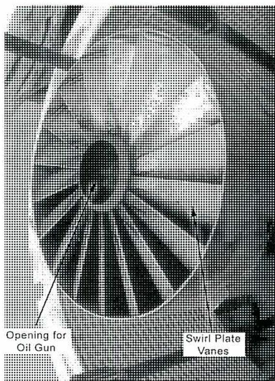Figure 14: A close-up photograph of a swirl plate assembly. It features a central circular opening for an oil gun and several curved, radial vanes (swirl plate vanes) that direct the flow of air or fuel to create a swirling motion. Labels with leader lines point to the 'Opening for Oil Gun' and the 'Swirl Plate Vanes'.