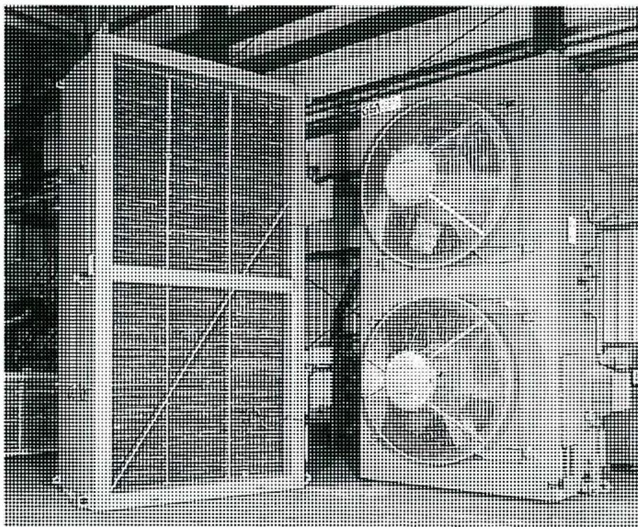 A black and white photograph of a large power transformer. The transformer has a prominent radiator section on its left side, consisting of several vertical panels. On the right side of the transformer, two large circular cooling fans are visible, mounted on the main tank. The entire unit is situated outdoors, with trees and a building in the background.