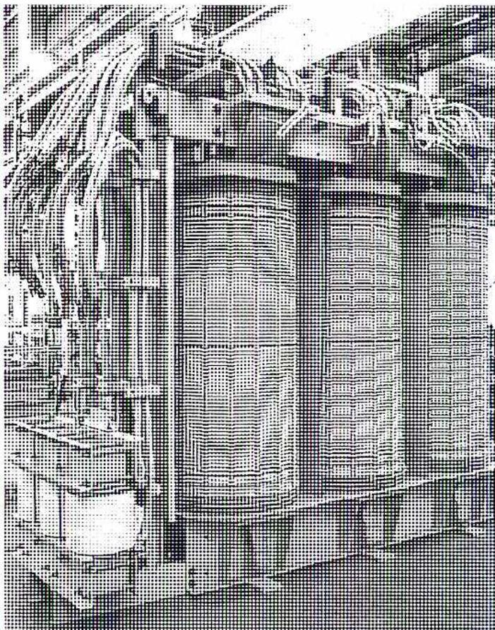 A black and white photograph showing three large, cylindrical transformer windings mounted on a core. The windings are arranged vertically, with the top one slightly offset to the left. The core is visible through the center of the windings, showing a rectangular shape. The windings are made of many layers of wire, and the top of each winding has several wires extending outwards.