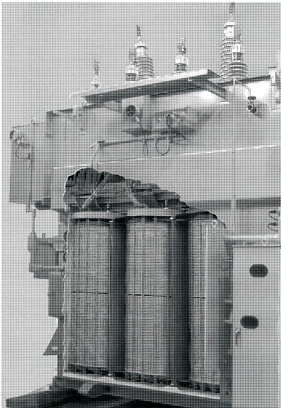 A black and white photograph of a large, industrial transformer enclosure. The enclosure is a tall, rectangular metal structure with a textured, possibly corrugated, surface. On top of the enclosure, there are several large, cylindrical components, likely bushings or insulators, arranged in a row. The base of the enclosure is surrounded by a dark, possibly oil-filled area. The overall appearance is that of a heavy-duty electrical component.